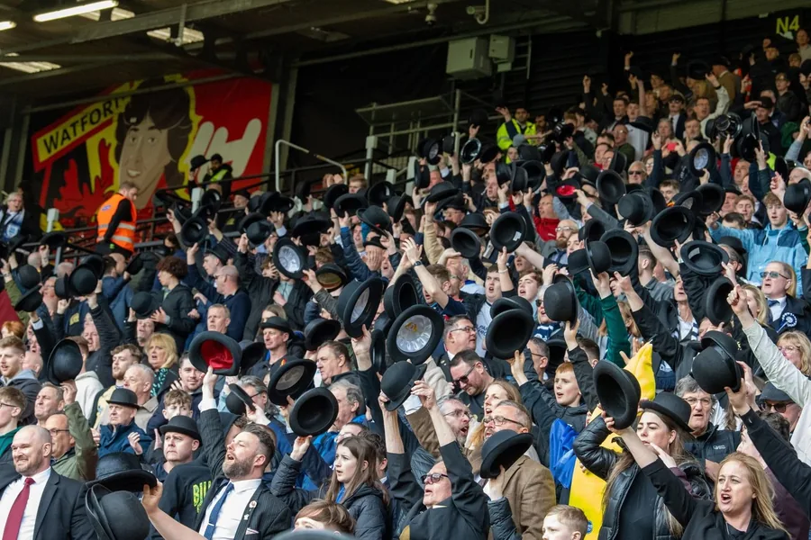 A wide stand full of Preston supporters raising bowler hats together.